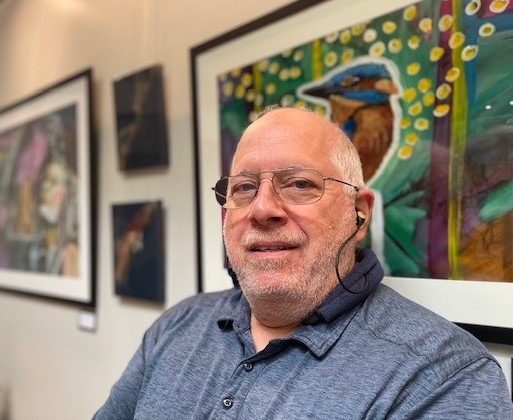 Tom Gisler, a man with gray hair and glasses, sitting in front of a painting