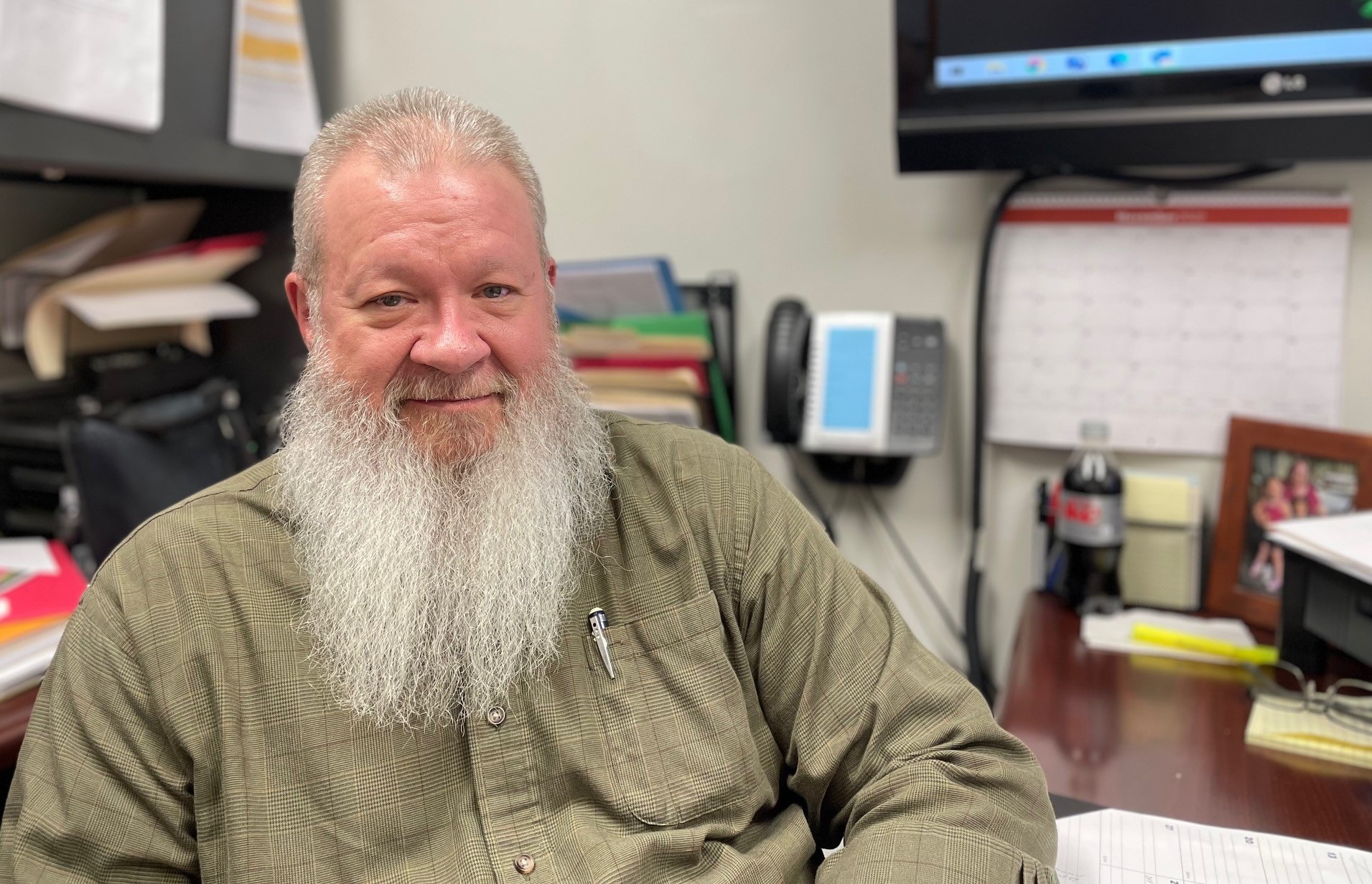 Bill Ryan sits at his desk. He is smiling and has a long white beard.