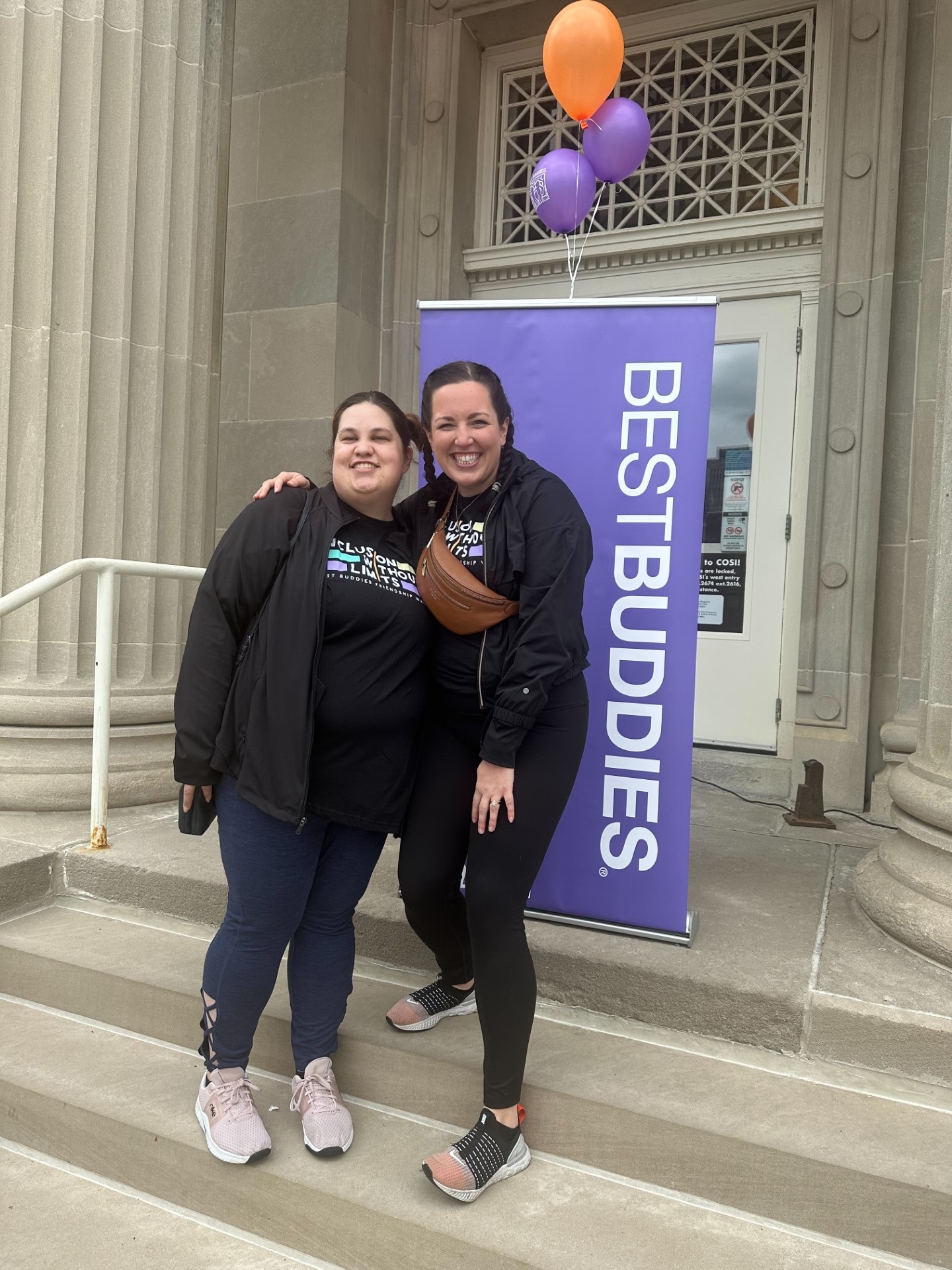 Two young women standing on front steps of a museum in front of a Best Buddies sign