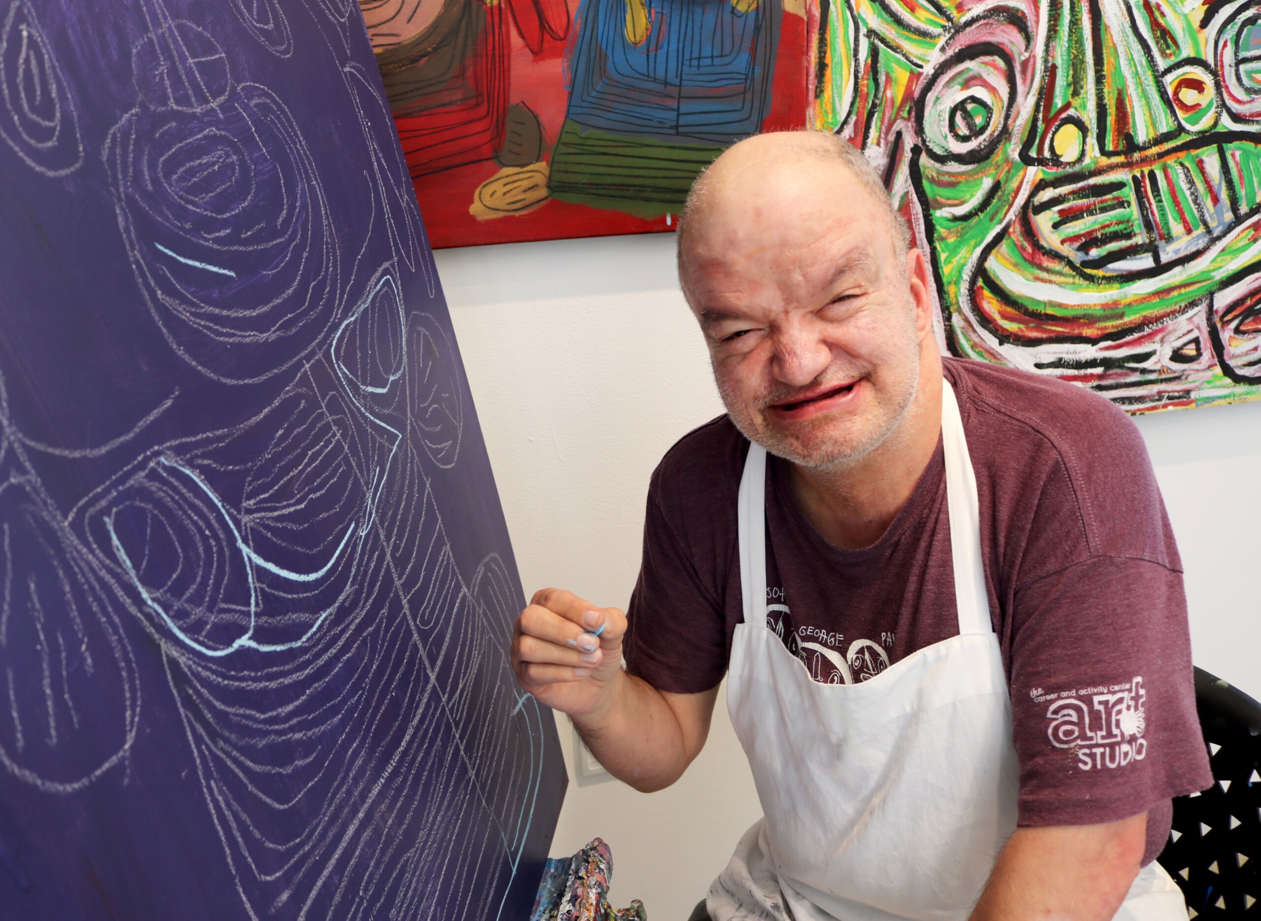 Wallace Peck, smiling and sitting in front of a dark blue canvas that he is sketching on.